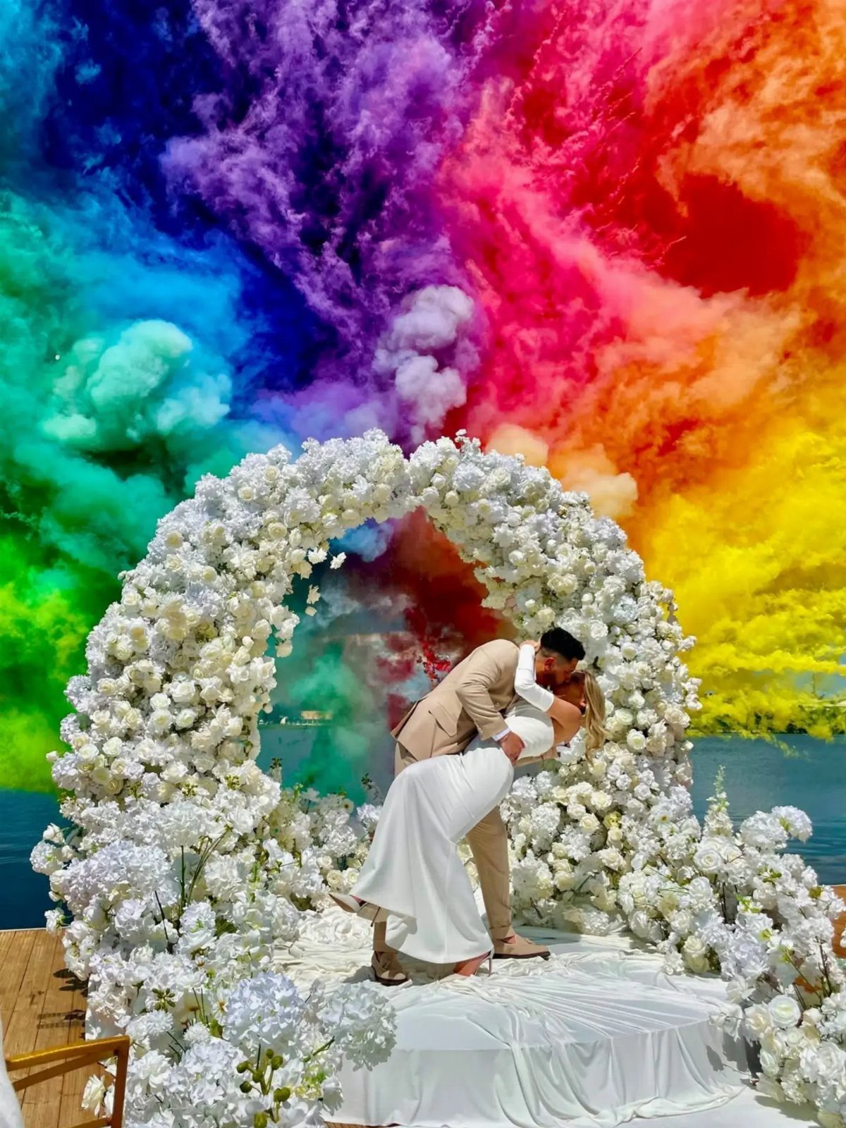 A bride and groom kissing under a white floral arch with colorful smoke in the background.