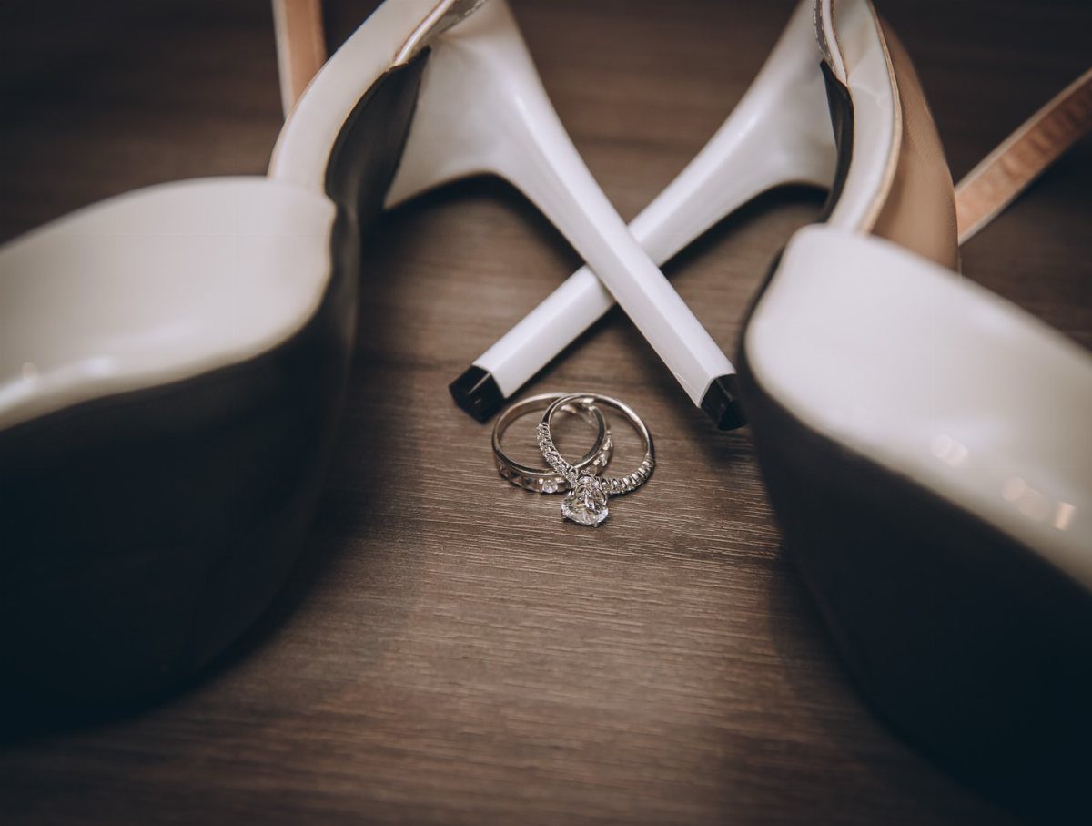 Two white high heels with crossed heels and diamond wedding rings on dark wood.