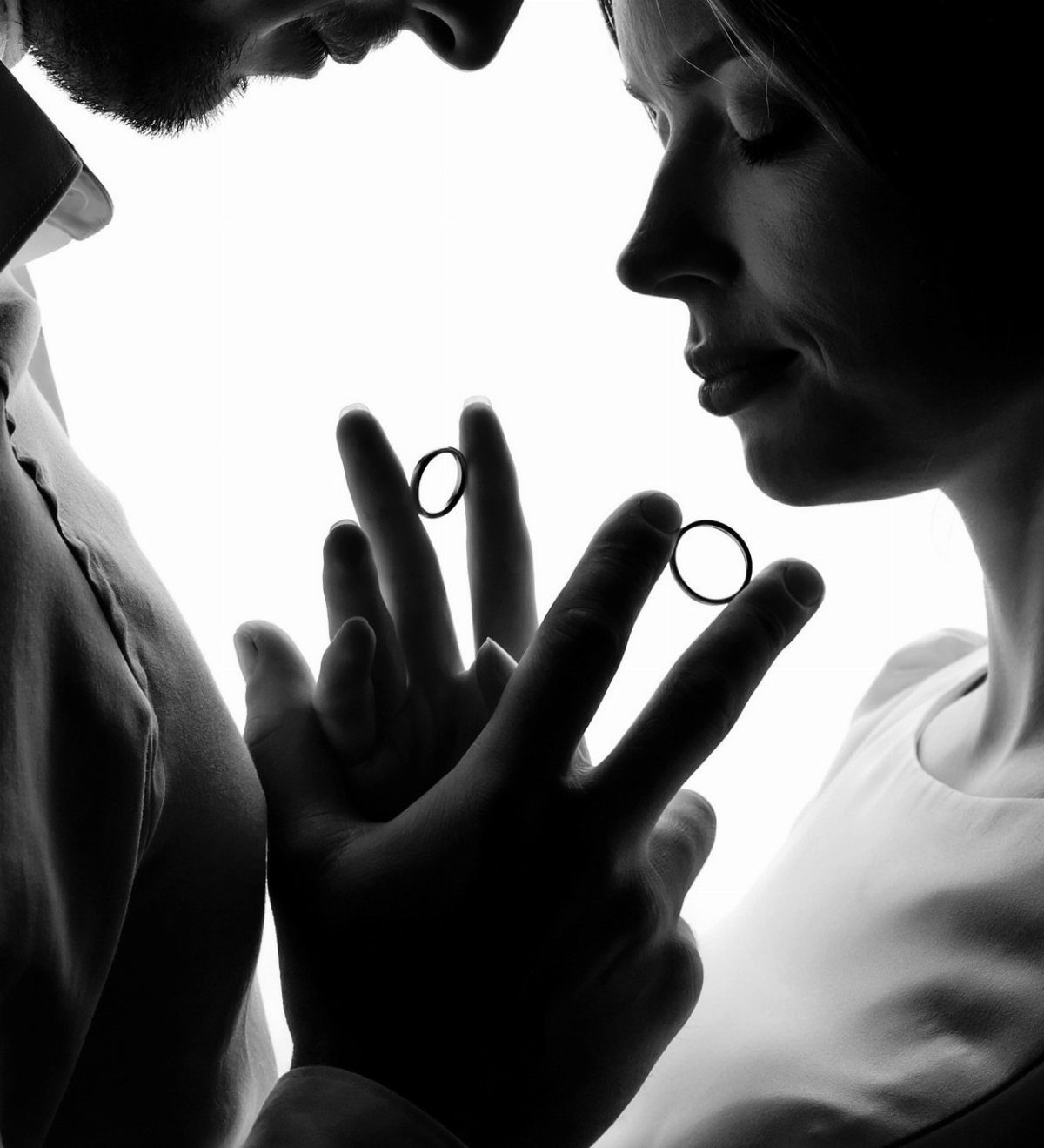 Silhouette of a couple holding wedding rings between their intertwined hands against a white background.