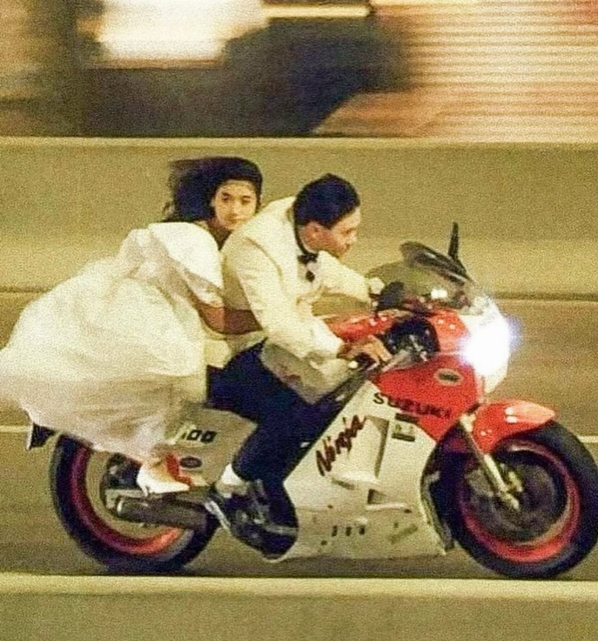 A bride and groom riding a red and white Suzuki Ninja motorcycle at night.