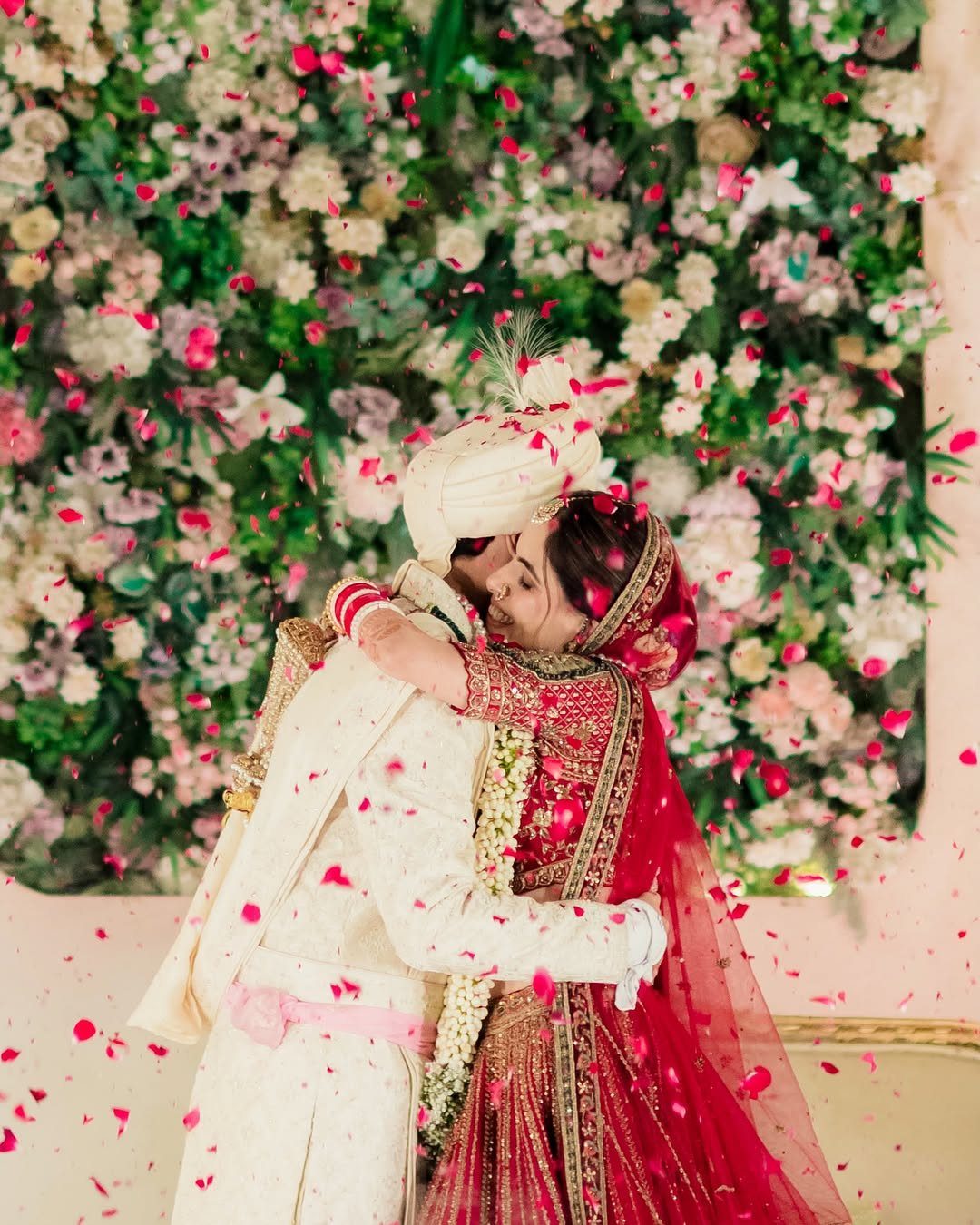 A bride and groom embracing warmly amidst falling pink flower petals against a floral backdrop.