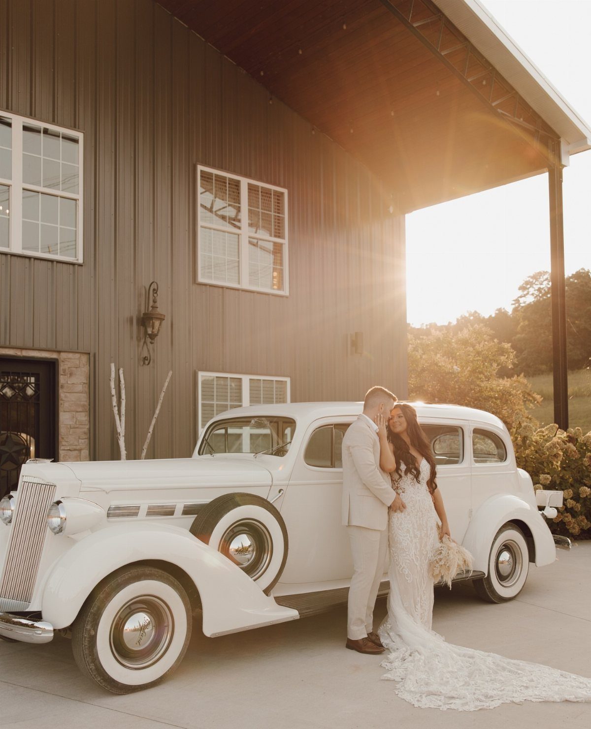 A bride and groom embrace beside a white vintage car in front of a barn.