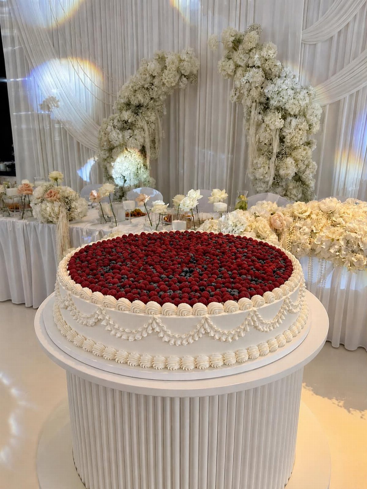 A white frosted cake topped with a thick layer of fresh red raspberries on a pedestal.