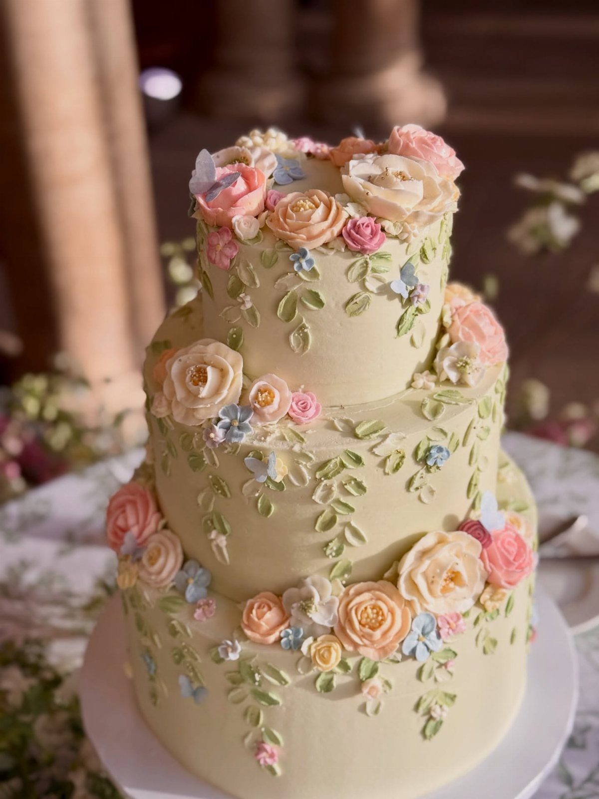 A three-tier pale yellow wedding cake decorated with colorful buttercream flowers and green leaves.
