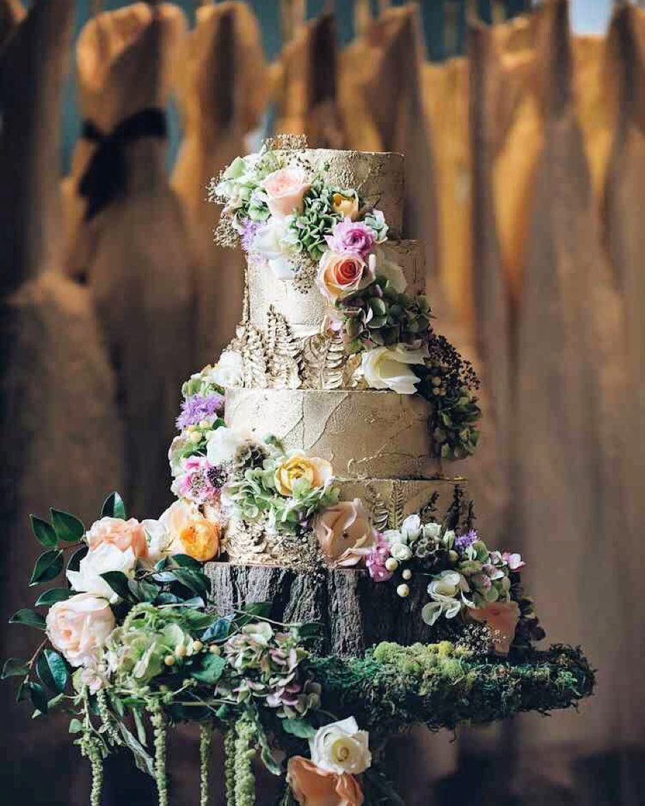 A tiered wedding cake decorated with pastel flowers and greenery sitting on a rustic tree stump.