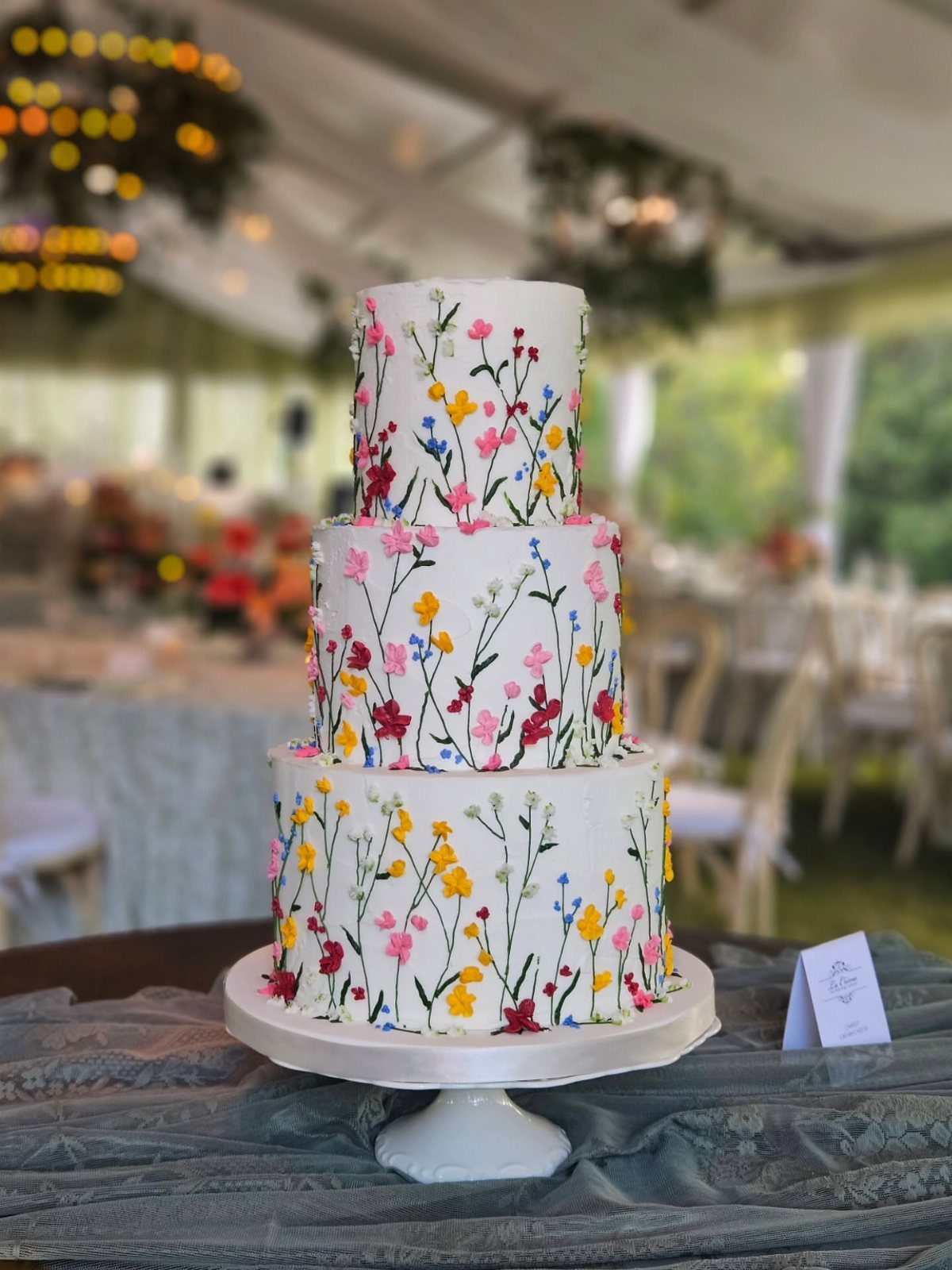 A three-tier white wedding cake decorated with colorful painted wildflowers on a white stand.
