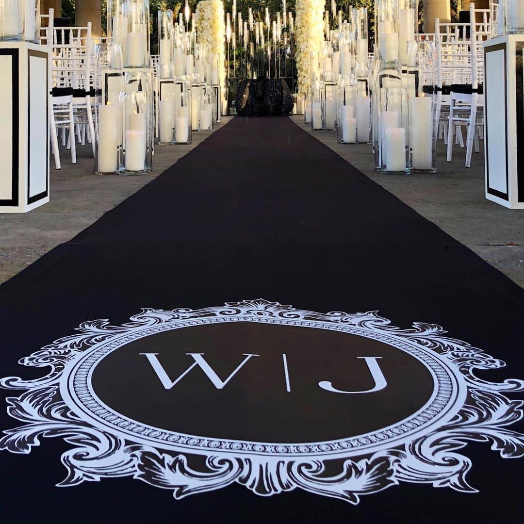 Black aisle runner with a white ornate monogram crest leading toward a wedding ceremony setup.