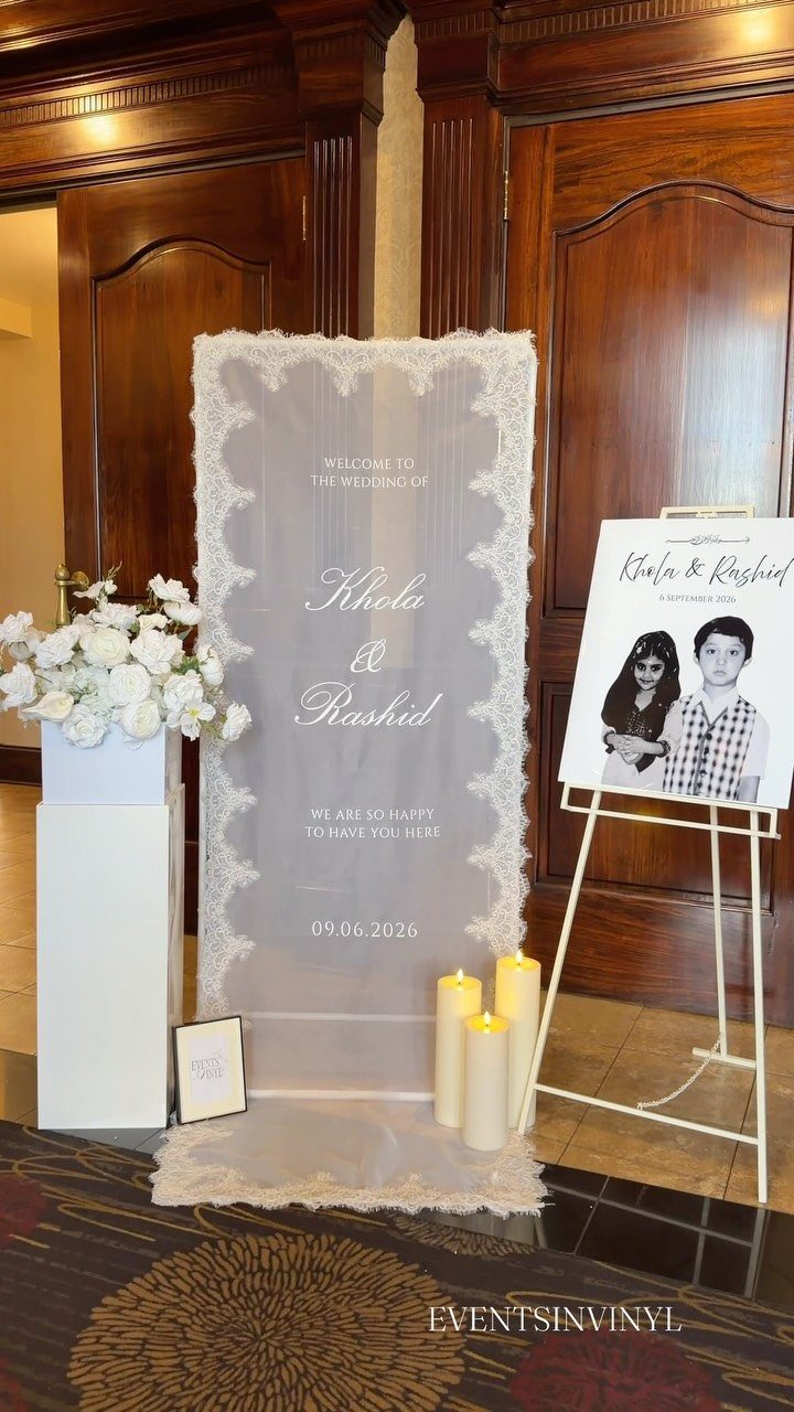 White wedding welcome signs on an easel and lace fabric stand near white flowers and candles.