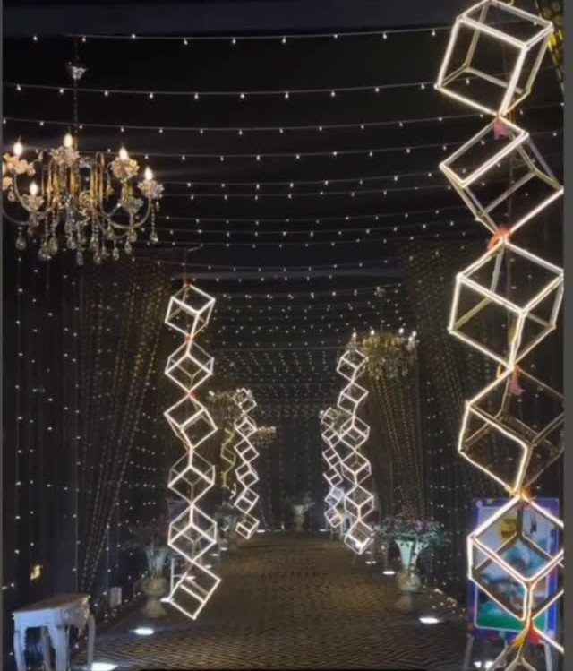 Glowing gold geometric wire cubes hanging above a dark walkway decorated with fairy lights and chandeliers.