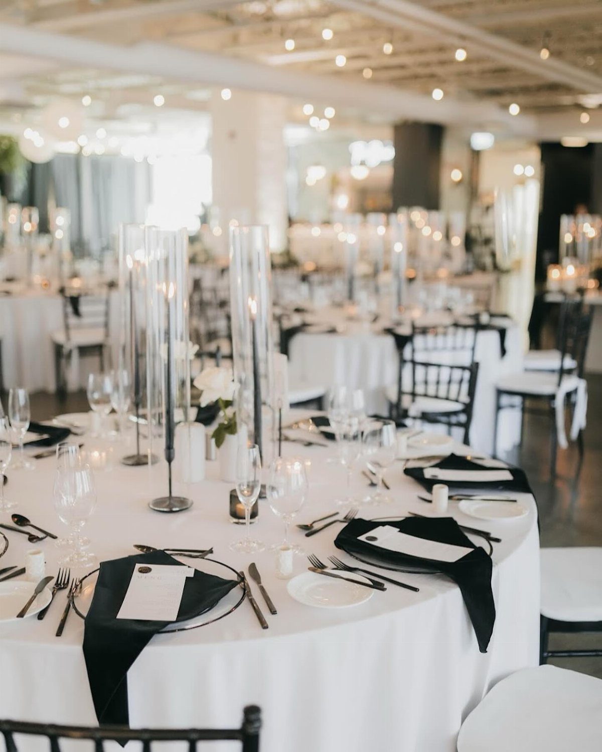 Formal wedding tables set with white tablecloths, black napkins, and tall glass candle holders.