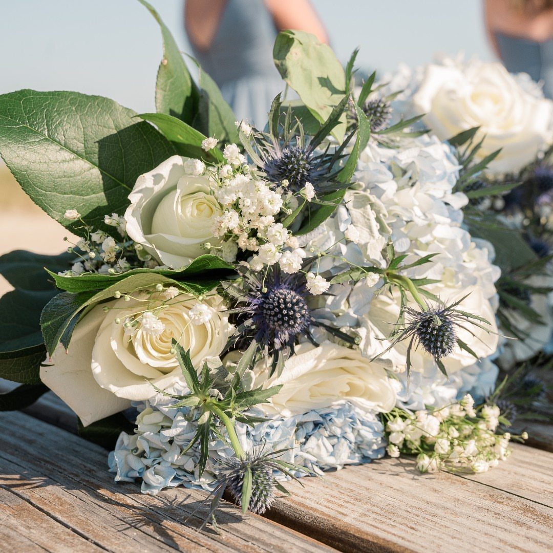 A wedding bouquet with white roses, blue hydrangeas, and blue sea holly thistles on wood.