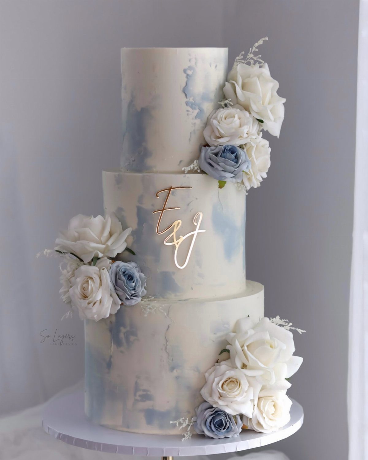 A three-tier white wedding cake decorated with light blue watercolor frosting and white and blue roses.