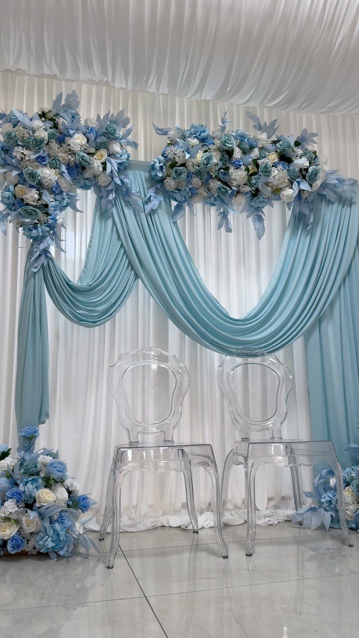 Blue and white floral arrangements draped over light blue fabric against a white backdrop with two clear chairs.