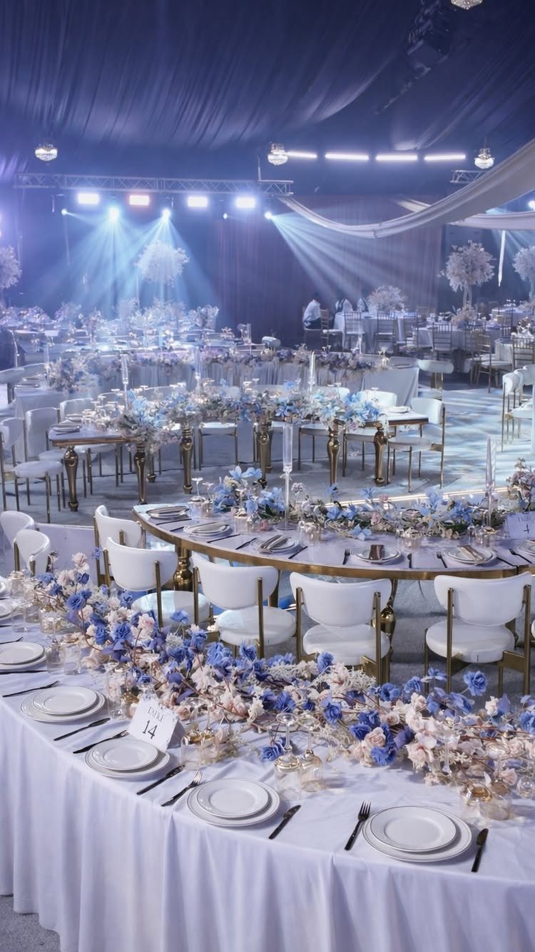 Wedding reception tables decorated with blue and white floral arrangements and gold chairs in a large hall.