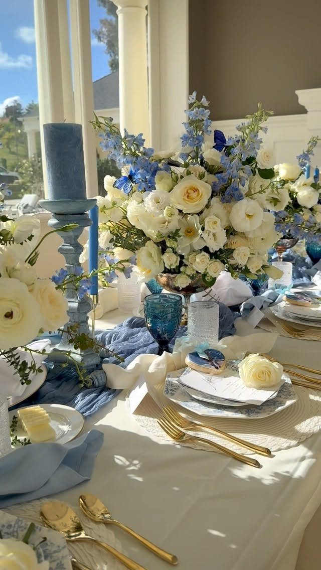 Wedding table setting with white roses, blue flowers, gold cutlery, and a dusty blue fabric runner.