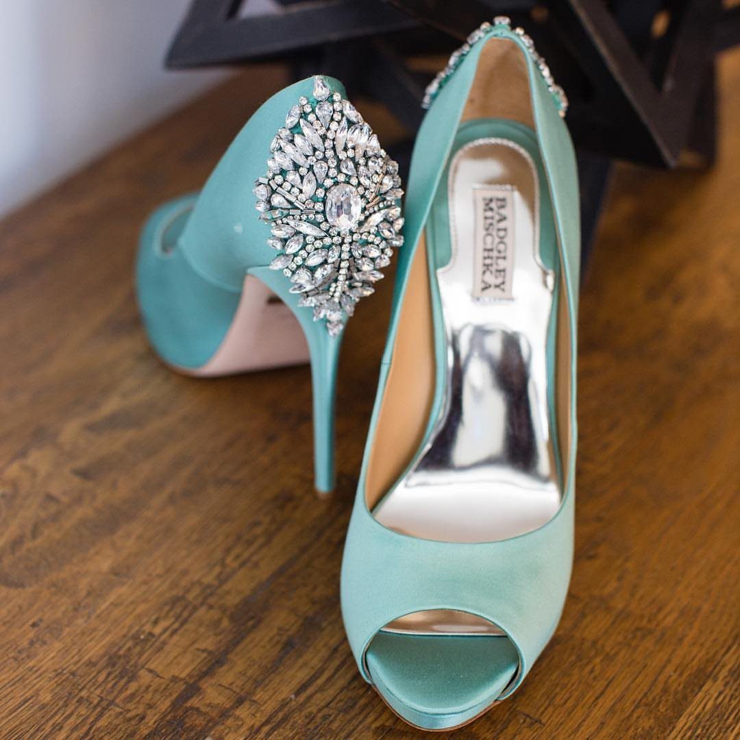 Teal satin peep-toe high heels with a large rhinestone embellishment on the heel sitting on wood.