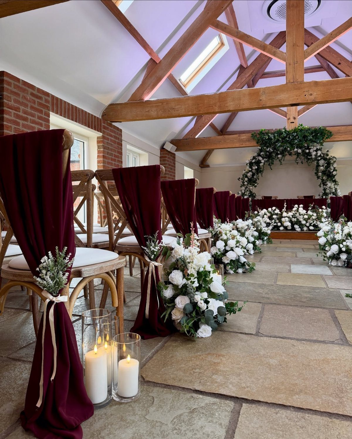 Wooden chairs with burgundy fabric sashes and white flowers lining a stone aisle.