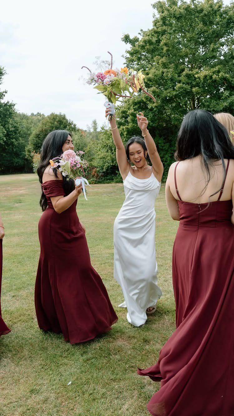 Three women in long dresses, including two in burgundy, standing on a green lawn.