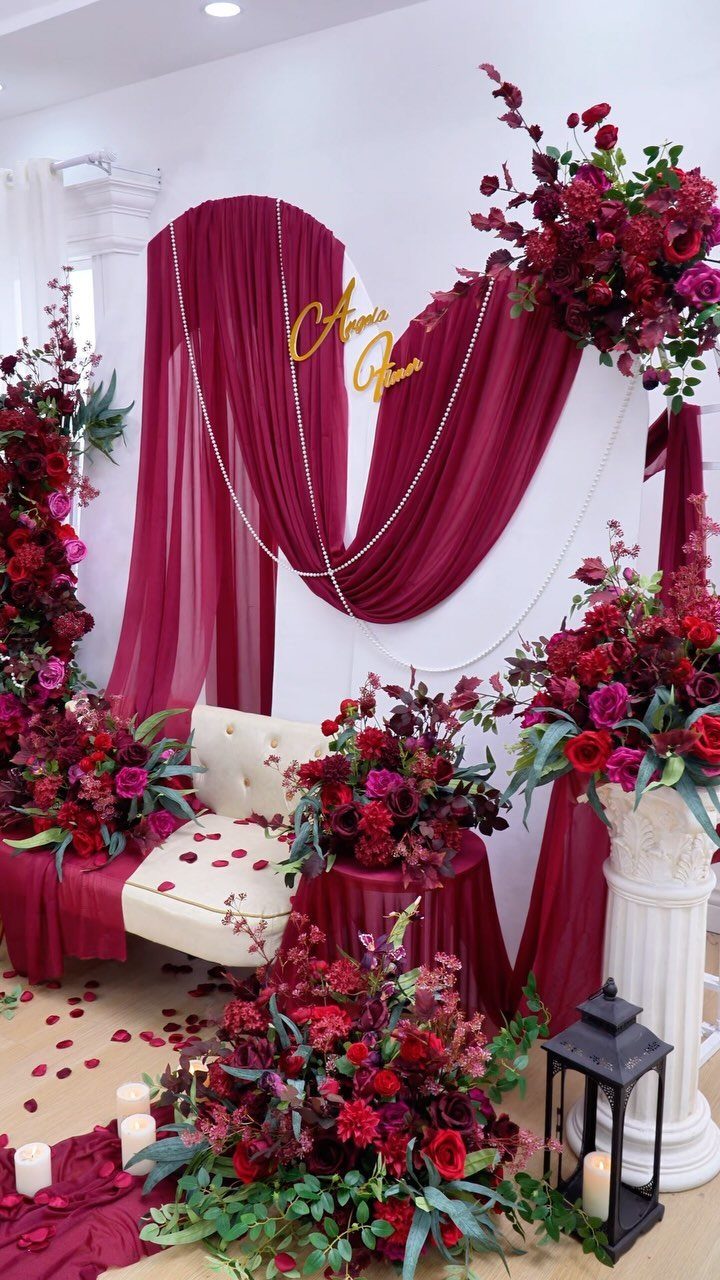 Burgundy fabric draping and large floral arrangements surrounding a white tufted bench in a wedding setting.