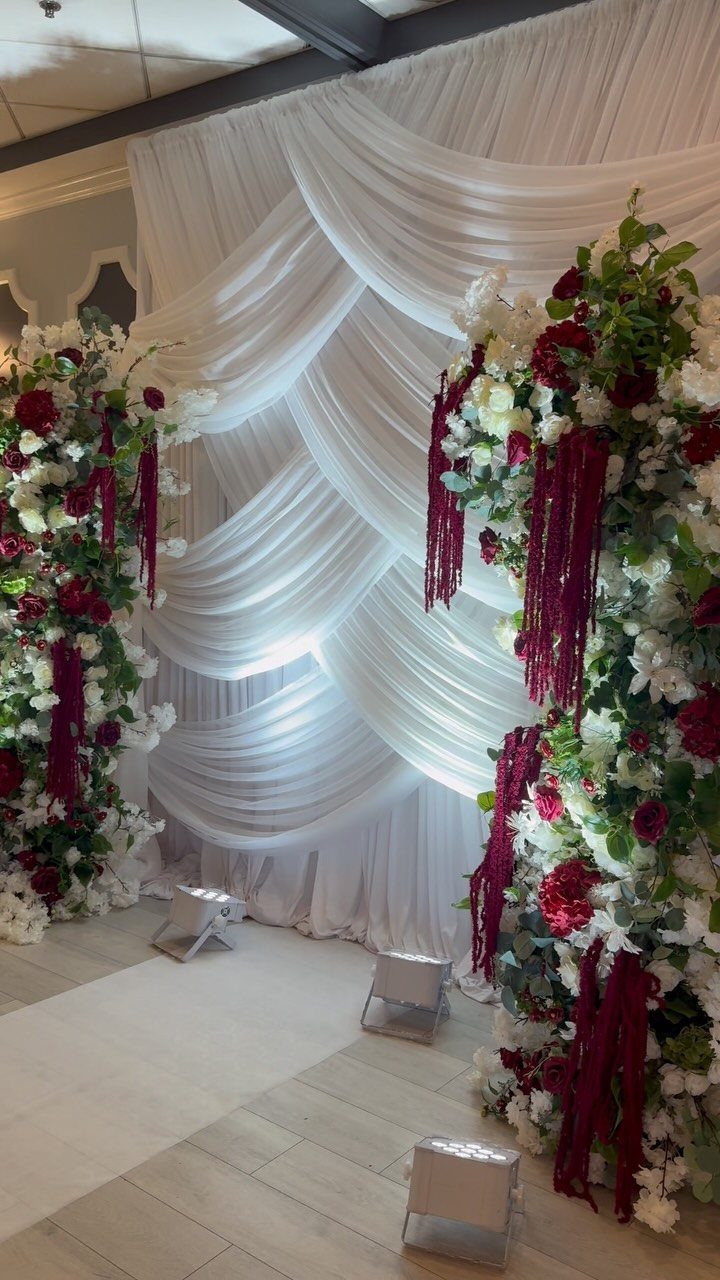 Large burgundy and white floral arrangements stand before a white draped fabric wedding backdrop.