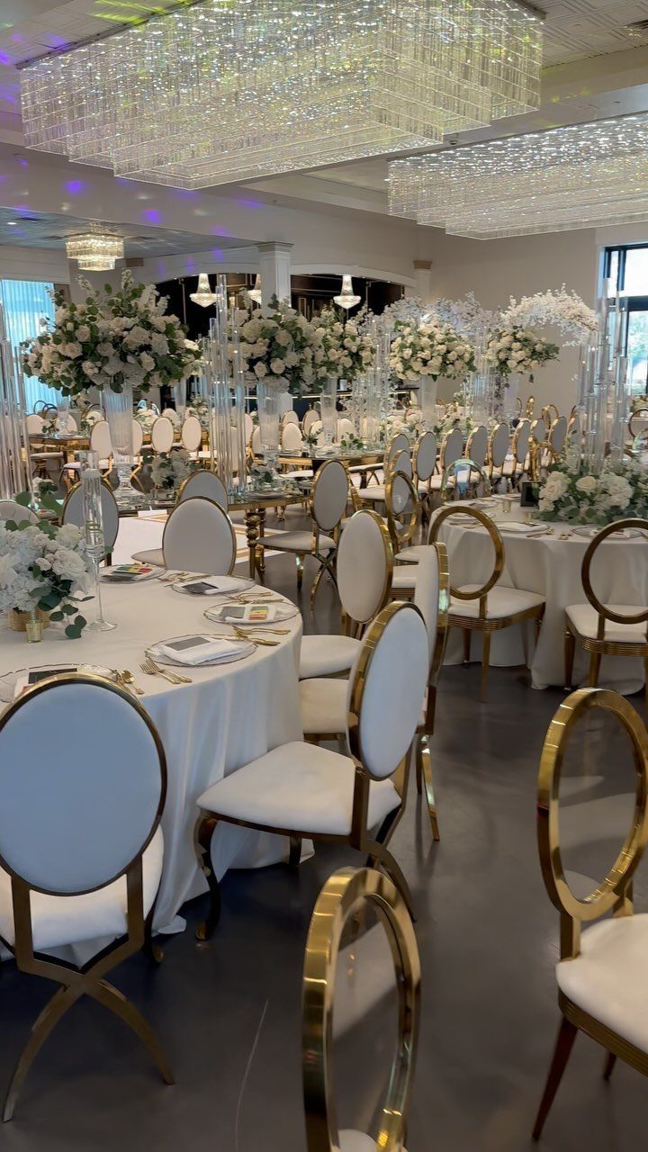 Elegant wedding reception tables with white floral centerpieces, gold chairs, and large crystal chandeliers.