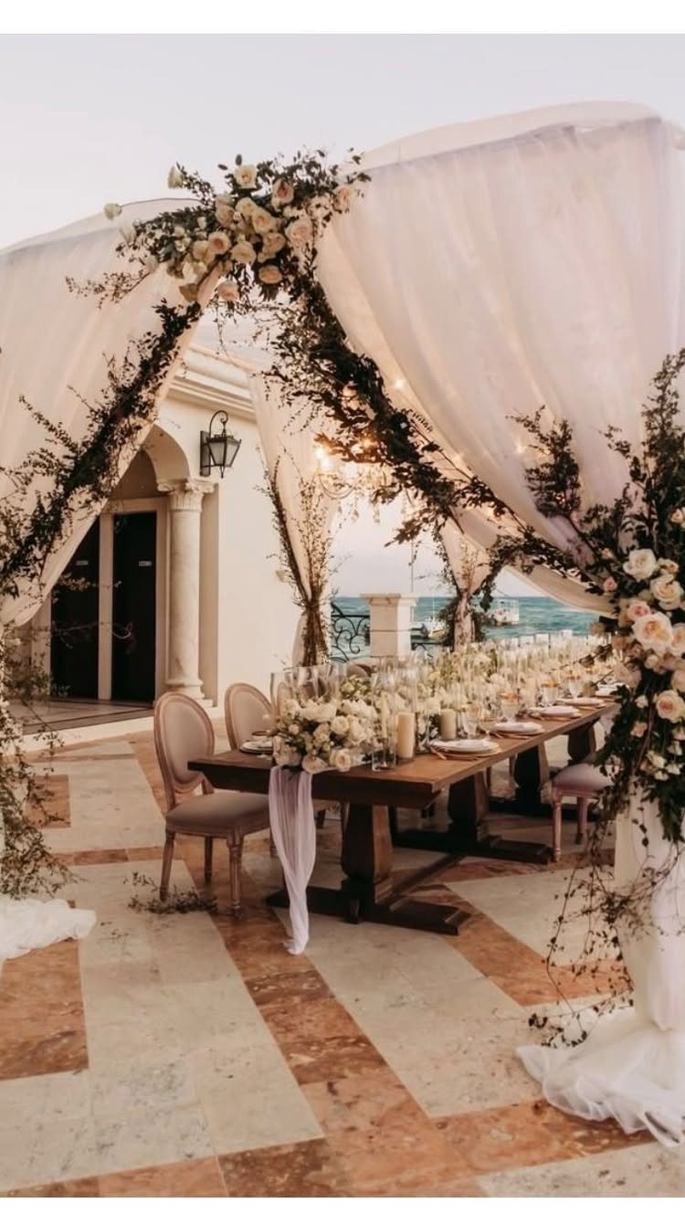 A long wooden wedding reception table decorated with white floral centerpieces and candles under a draped canopy.