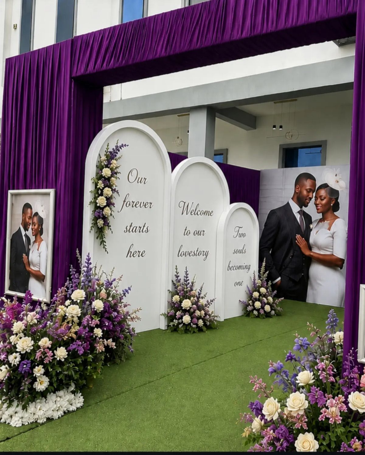 Purple fabric draping frames white arched backdrops decorated with flowers and text at a wedding entrance.