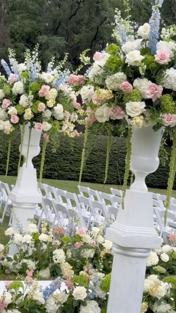 Large white urns filled with pink and white roses and blue flowers at an outdoor wedding.