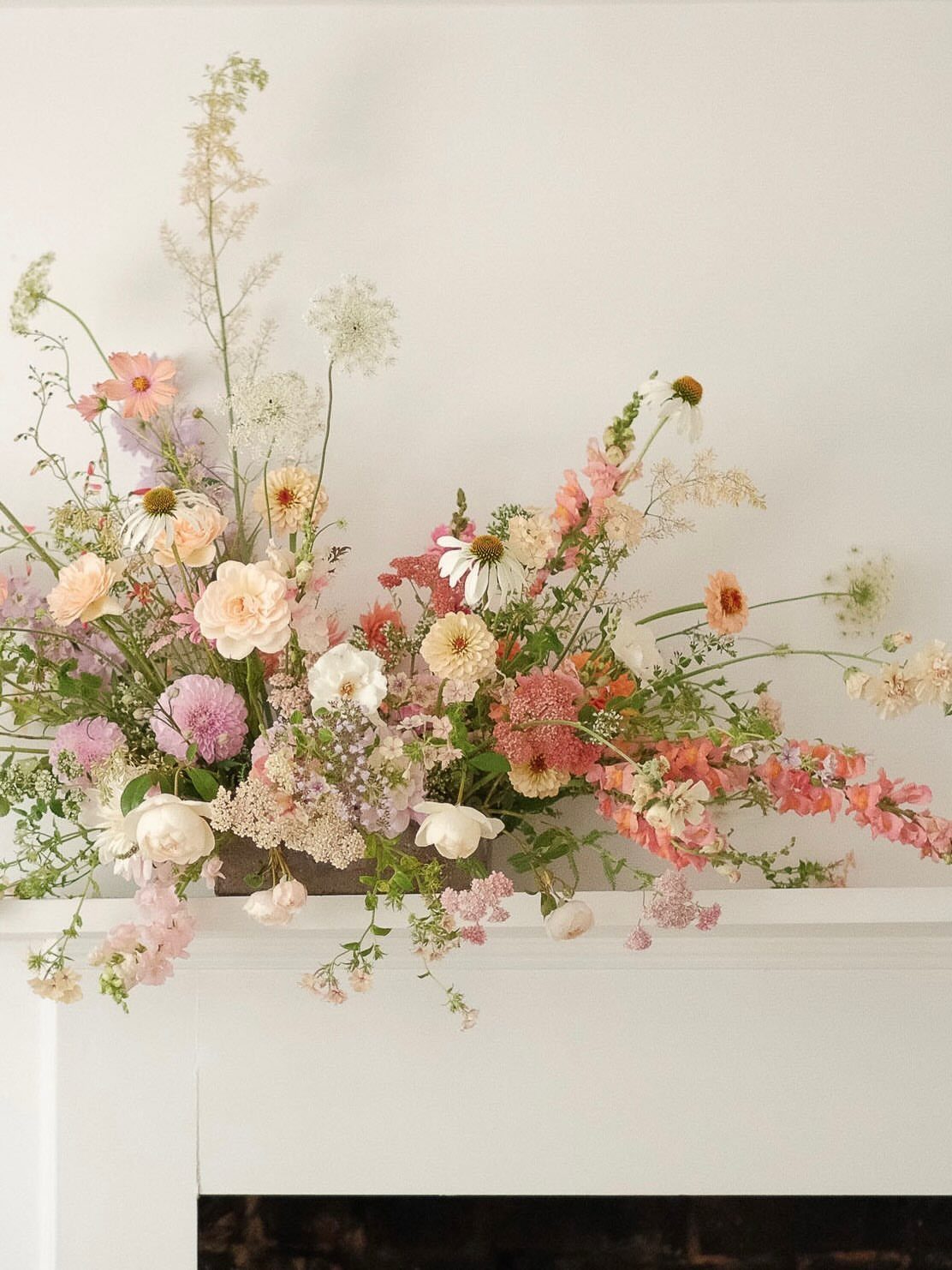 Wildflower arrangement with peach and white blooms resting on a white mantelpiece against a white wall.