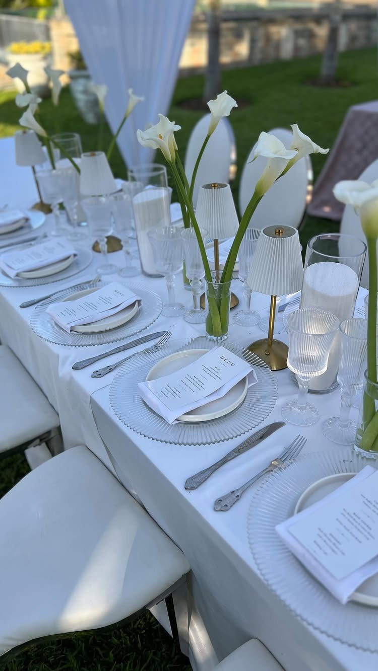 An elegant outdoor wedding table setting featuring white calla lilies, gold lamps, and fine glassware.