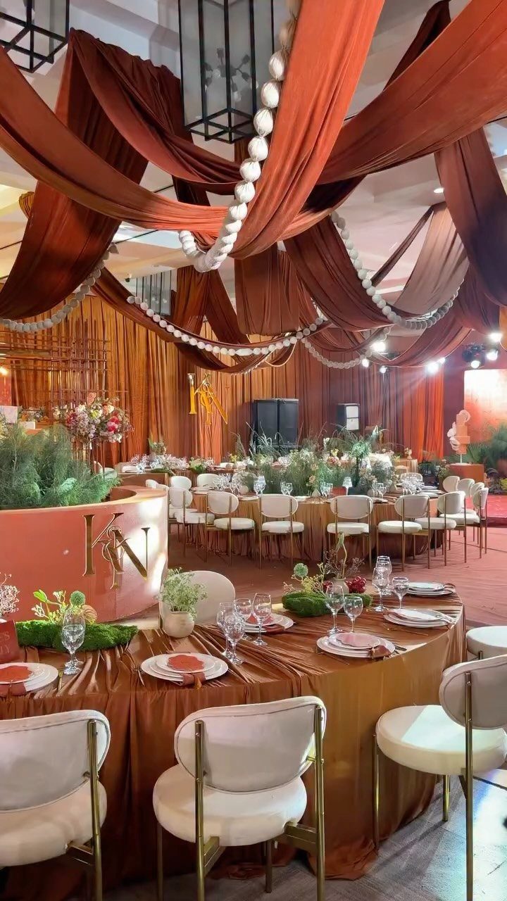 Terracotta fabric draping and pearl garlands decorate a wedding reception hall with elegant table settings.