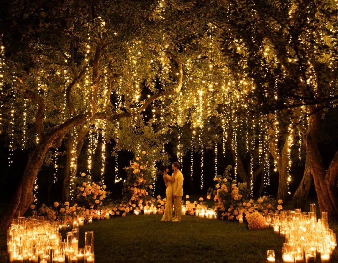 A couple stands under trees decorated with hanging fairy lights and surrounded by glowing candles and flowers.