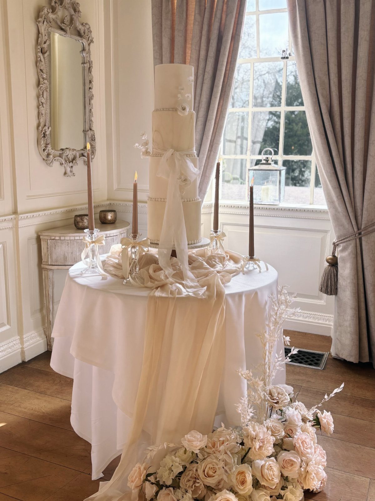 A tall cream wedding cake on a white draped table with candles and roses.