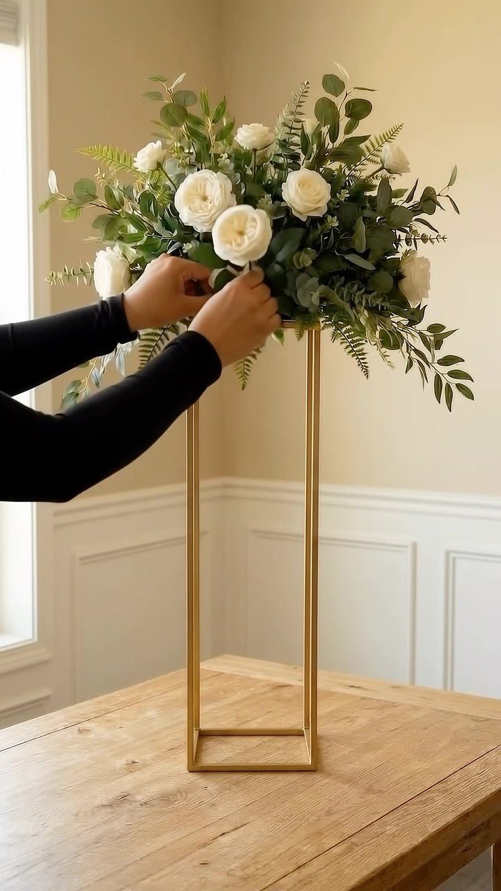 Hands arranging white roses and greenery into a tall gold metal pedestal stand.