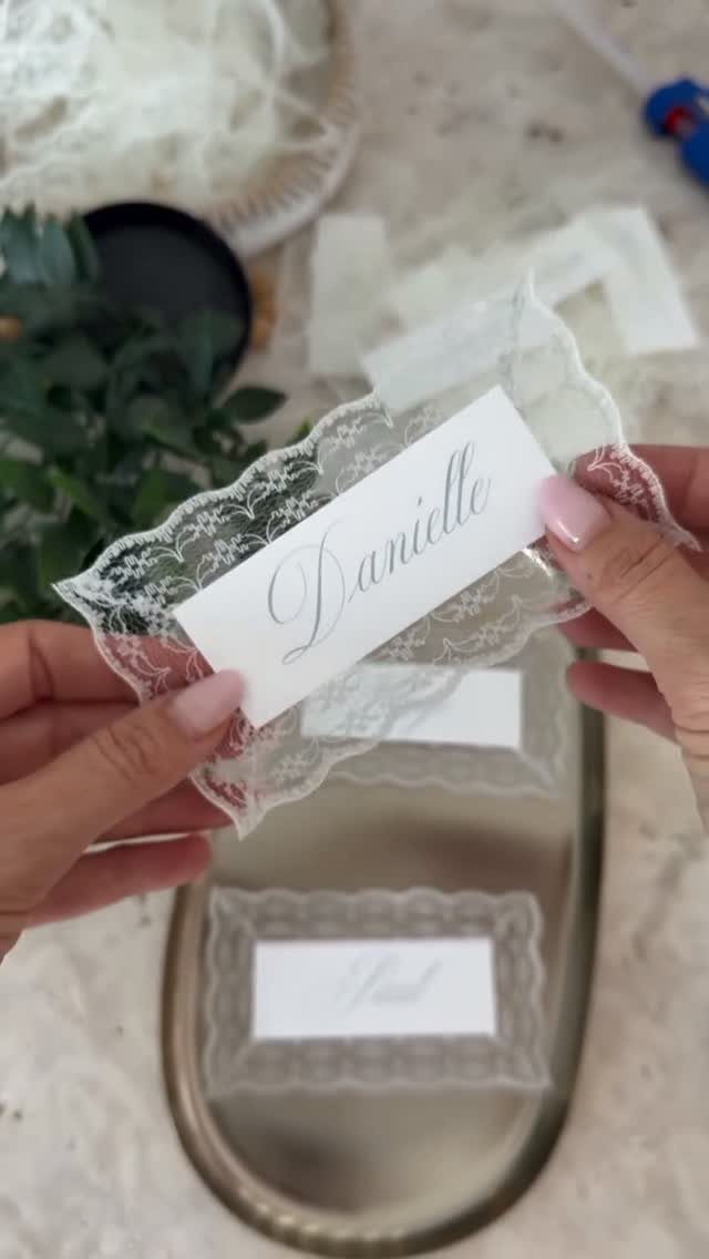 Hands holding a white lace-edged placecard with the name Danielle printed in elegant script.