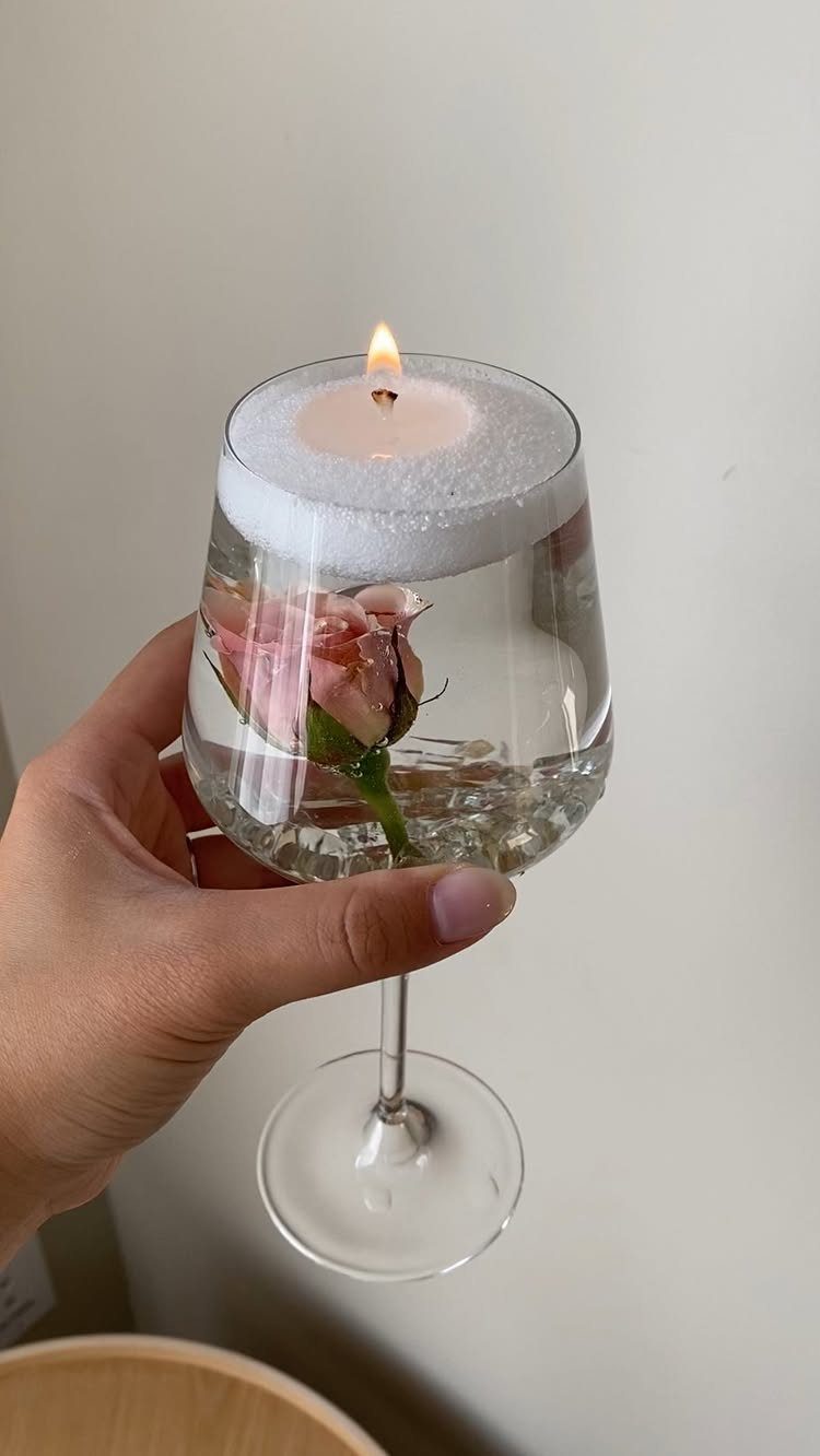 A hand holding a wine glass containing a rose and a floating lit candle.