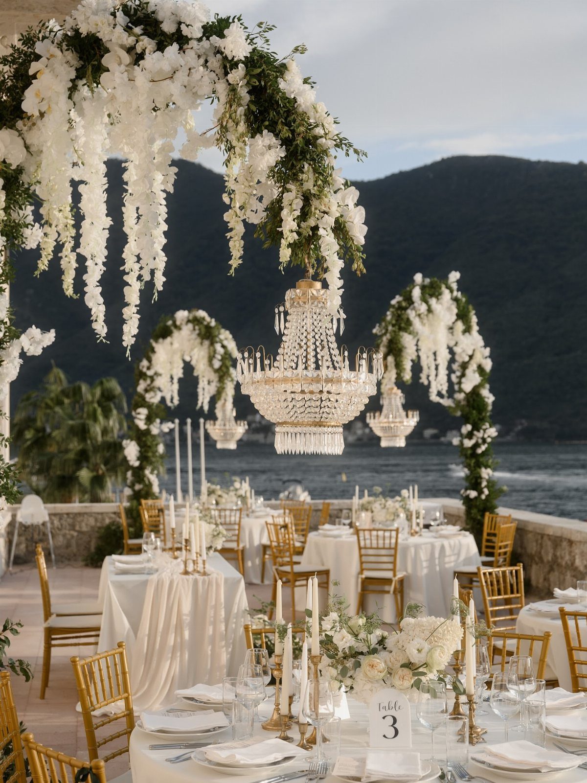Crystal chandeliers hanging over white wedding tables on an outdoor terrace by the sea.