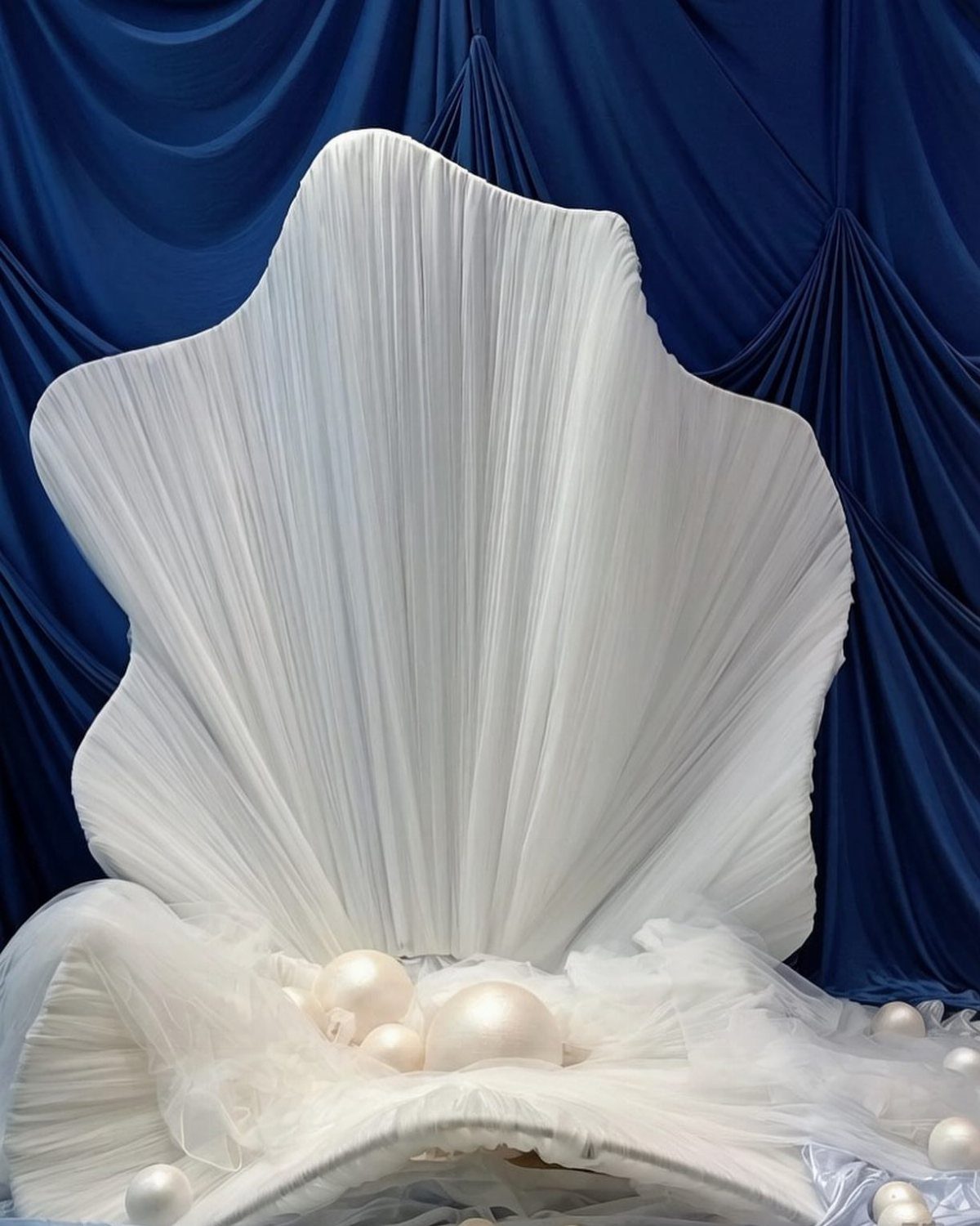 A white pleated fabric chair shaped like a large seashell against blue drapes.