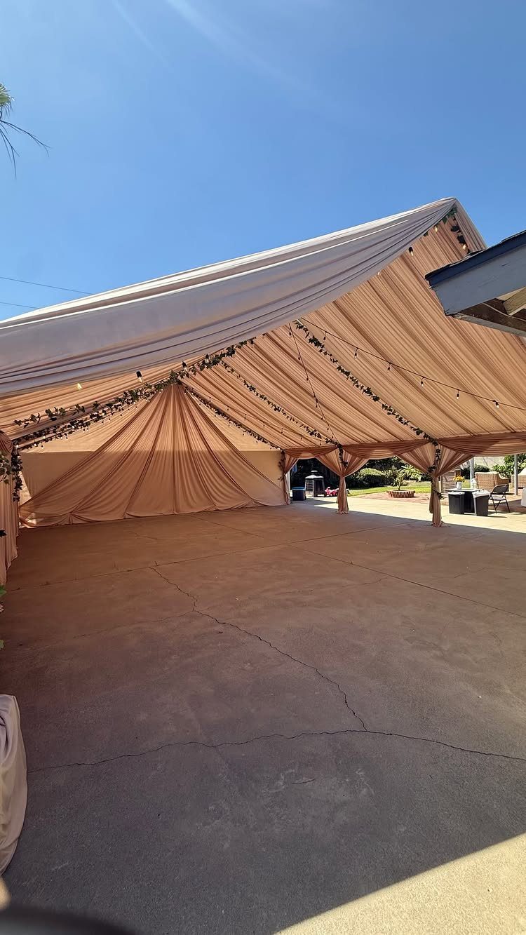 Beige fabric draping and string lights inside a large outdoor event tent under a blue sky.