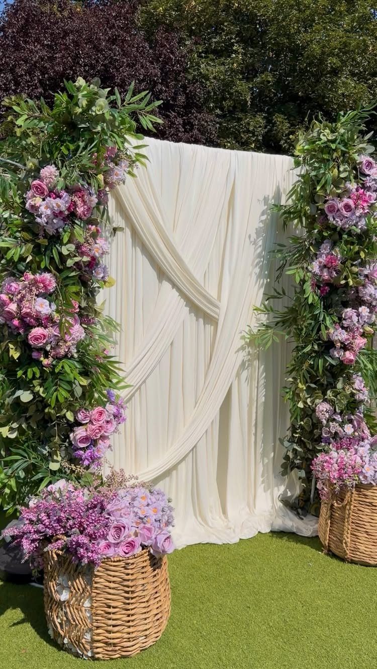 Pink and purple floral installations framing a white draped fabric backdrop on green grass.