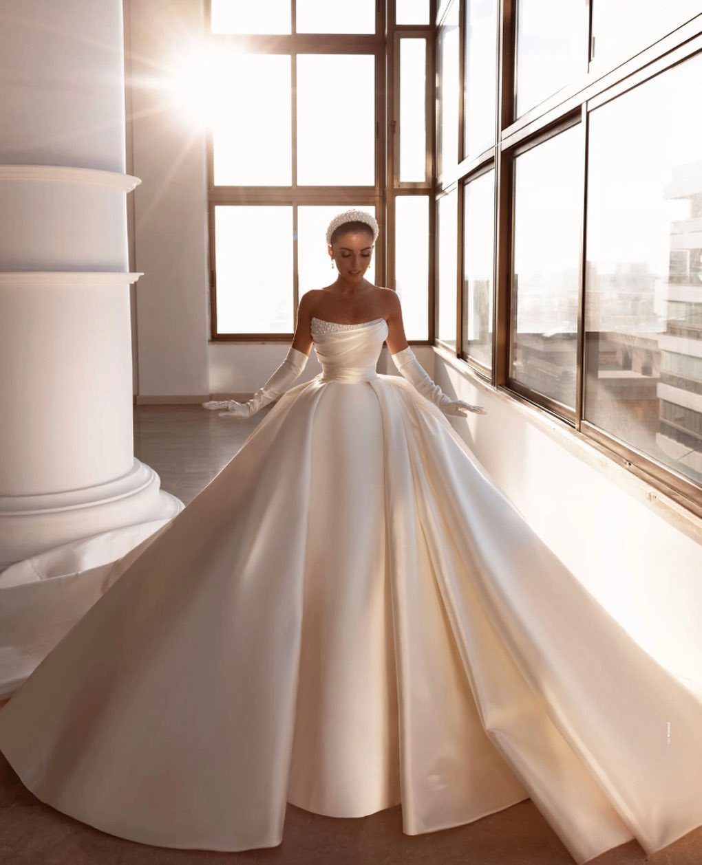 Woman wearing a voluminous ivory satin princess ball gown standing in a sunlit room by large windows.