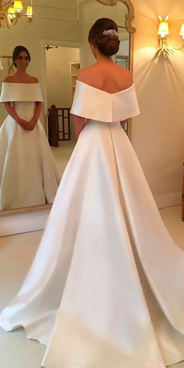 7 Wedding Dresses for Winter Brides That Balance Warmth and Style 3 A woman wearing a white satin off-the-shoulder A-line wedding dress standing in front of a mirror.