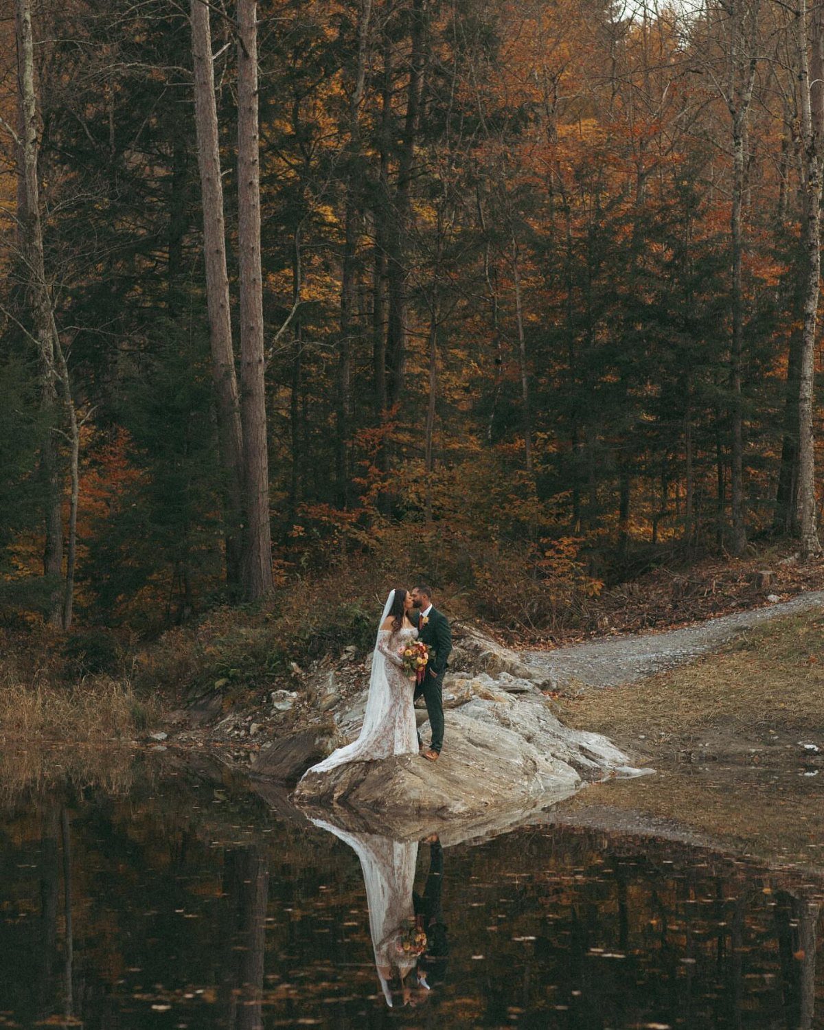 A bride and groom pose on a rock by a lake amidst an autumn forest.