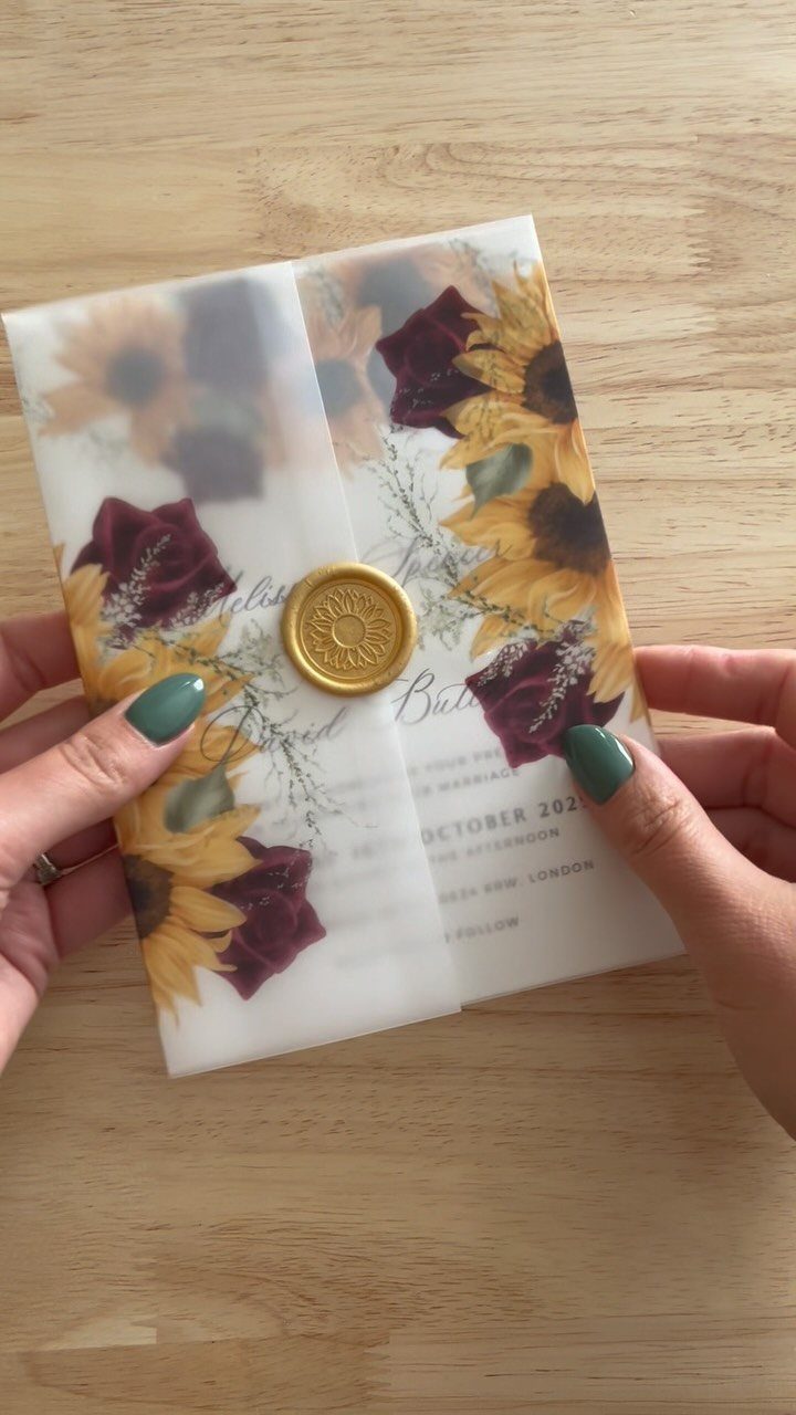Hands holding a vellum wedding invitation with sunflower and rose illustrations and a gold wax seal.