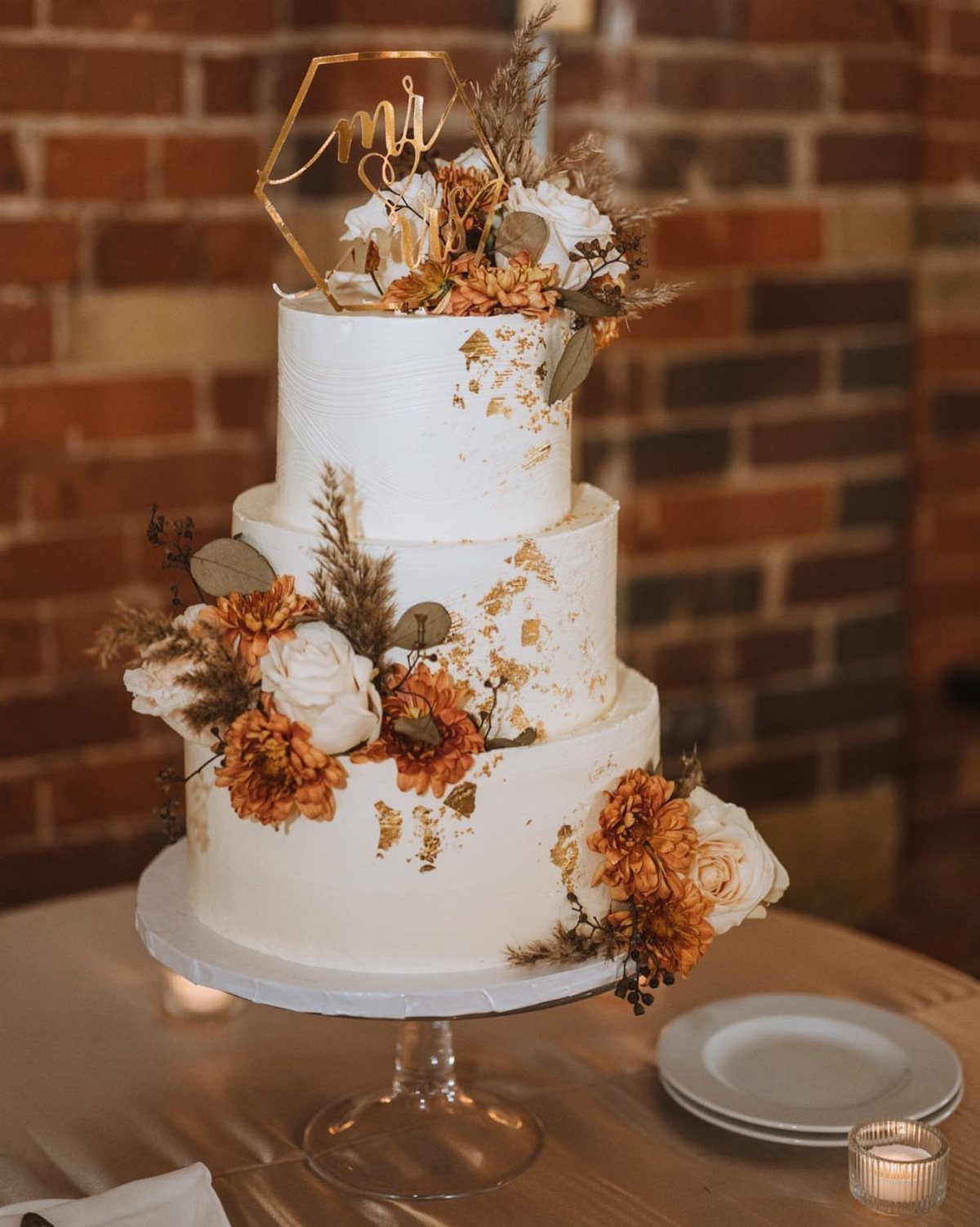 A three-tiered white wedding cake decorated with gold leaf and autumn flowers on a glass stand.