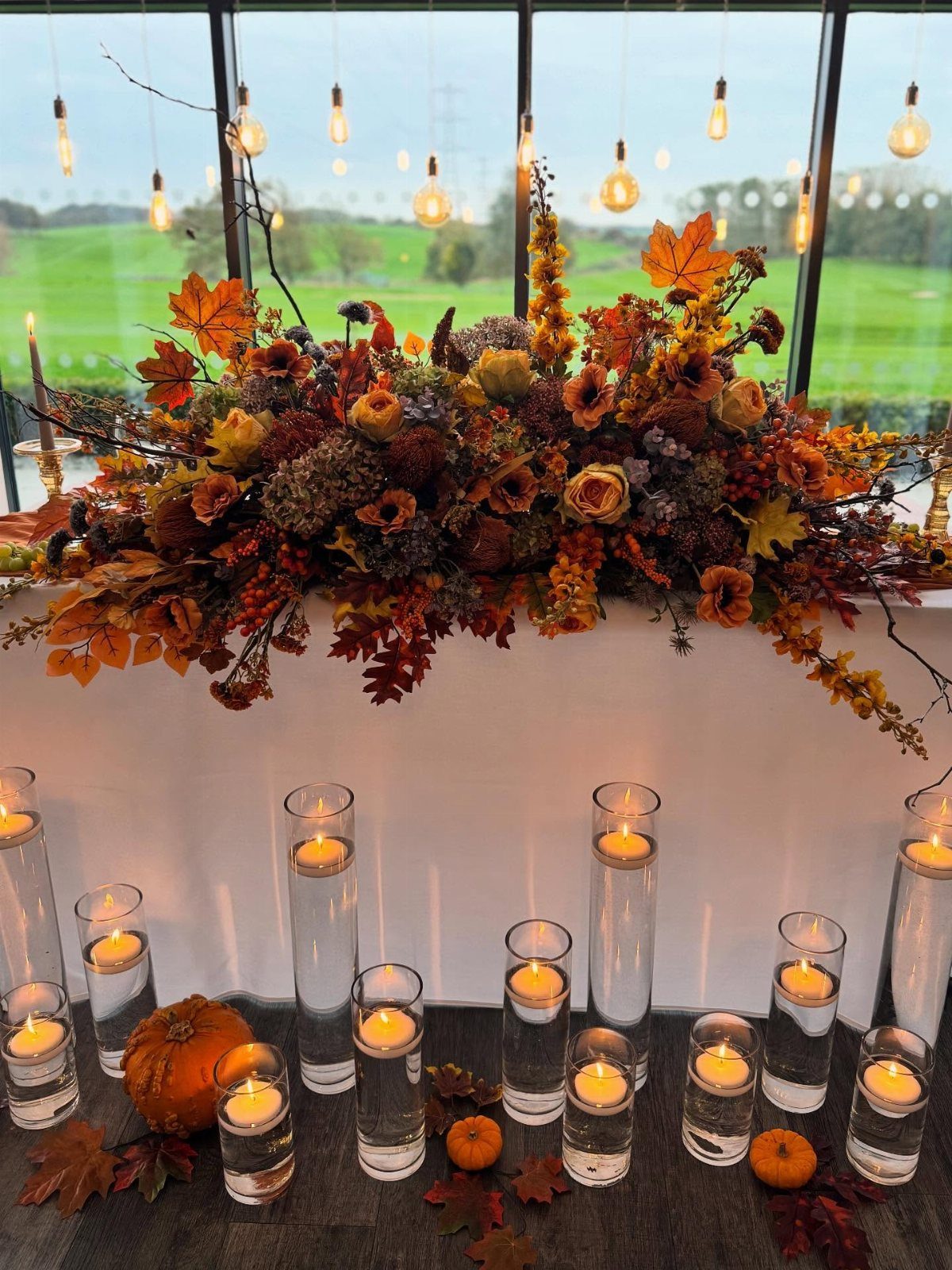 Autumnal floral arrangement with orange leaves and roses above floating candles in glass cylinders.