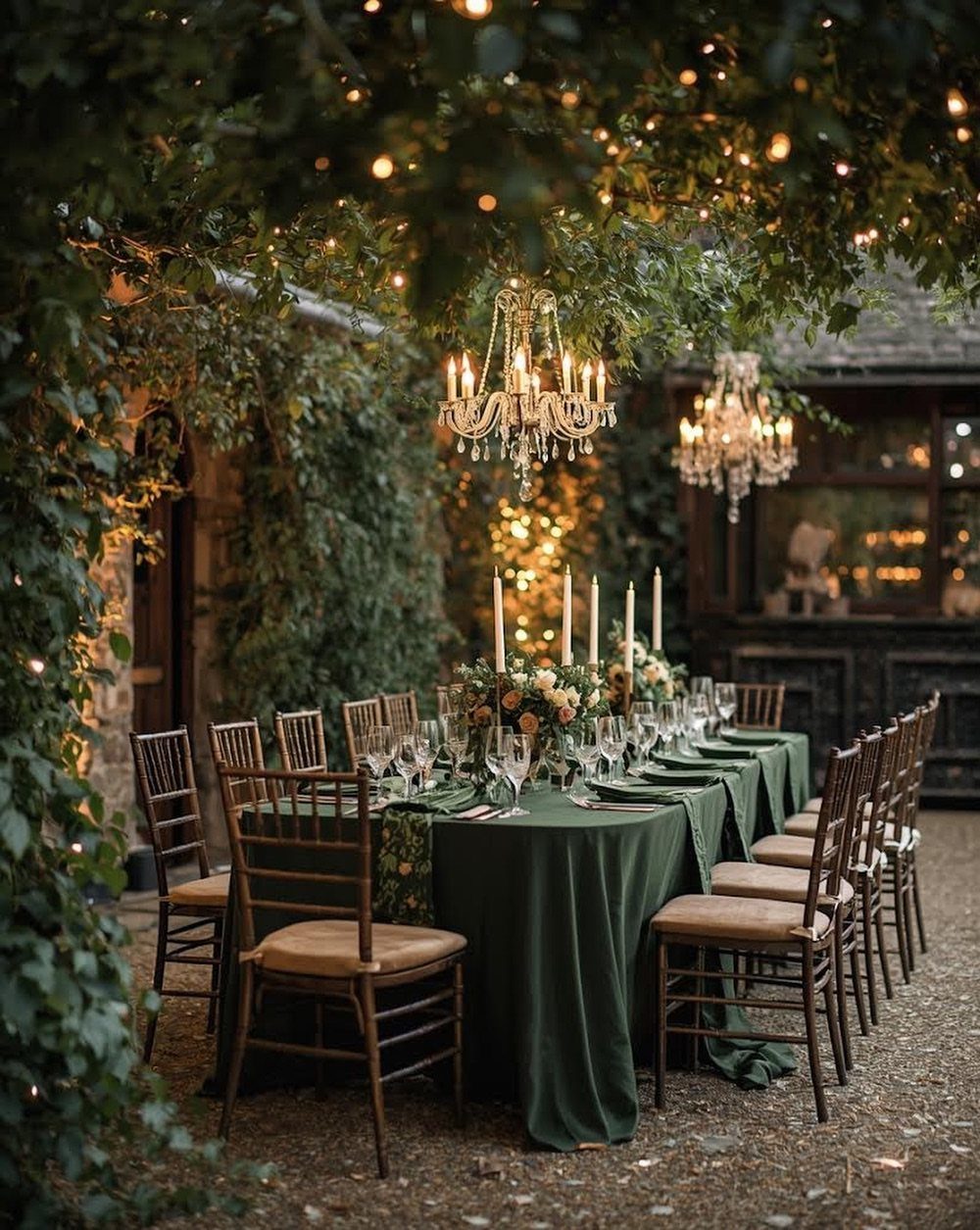 A long outdoor dining table set with green linens, candles, and crystal chandeliers under trees.