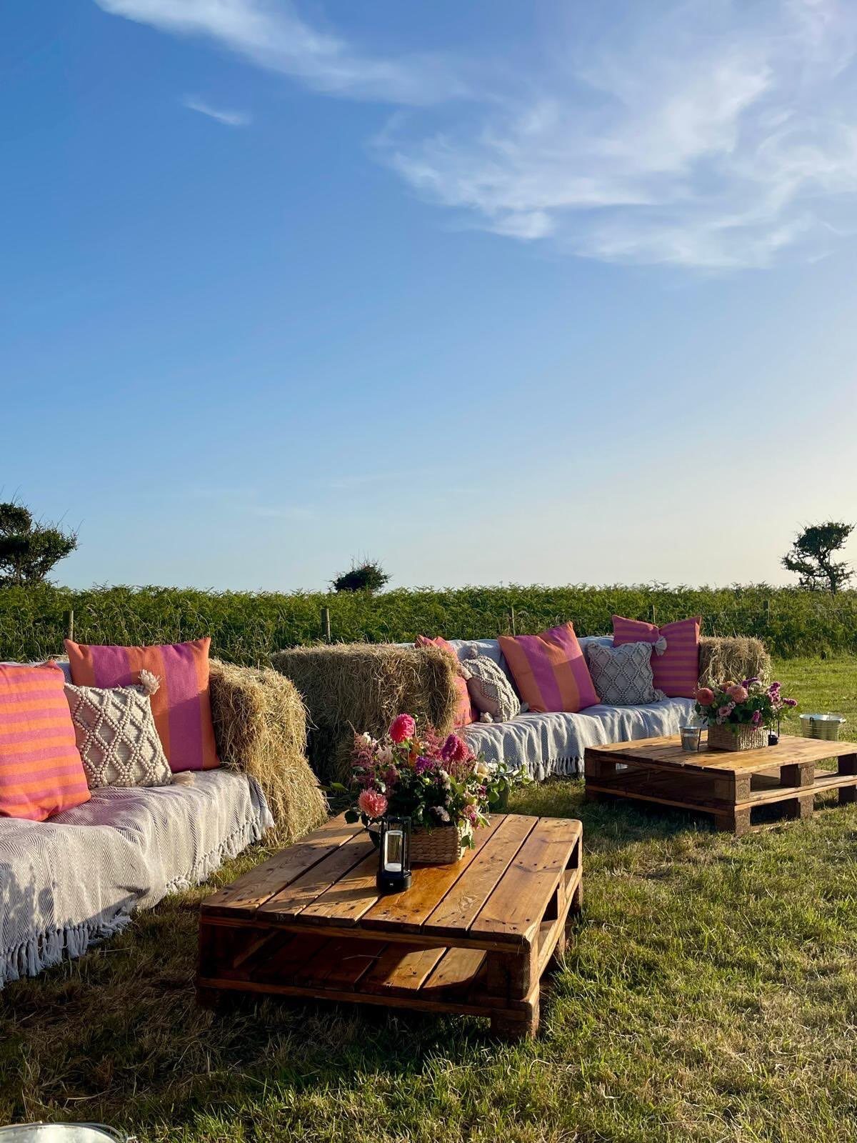 Hay bales with blankets and pink pillows arranged as outdoor seating with wooden pallet tables.