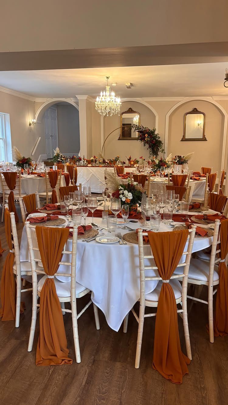 Wedding reception tables decorated with white linens, orange chair sashes, and autumnal floral centerpieces.
