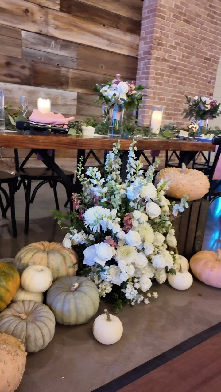 Assorted white and pale green pumpkins resting on the floor beside a large white floral arrangement.